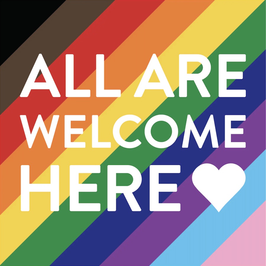 All are welcome here sign on a rainbow Pride flag background